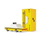 Small, geometric wooden taxi next to its yellow packaging.
