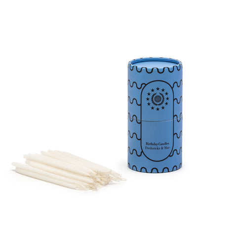 Image featuring a standing muted blue colored cylinder with black linear wave details along its sides along with the company name and logo, as the container for a set of small white birthday cake candle sticks laying down flat.