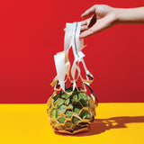 Against a yellow ground and red wall, a hand stretches from right, holding the handles of a net-style bag made of thin, flat, die-cut paper, which holds a head of lettuce. 