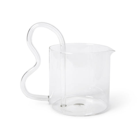 A clear glass pitcher with a short, wide, and round body. A thick, curved, bean-shaped glass handle is affixed near the base on both the exterior and interior, hovering over the edge of the pitcher.