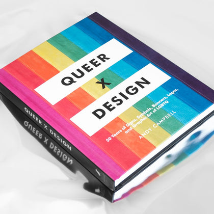 A book whose cover features the rainbow Pride Flag with text reading "Queer X Design",  lying on a silvery, reflective surface.