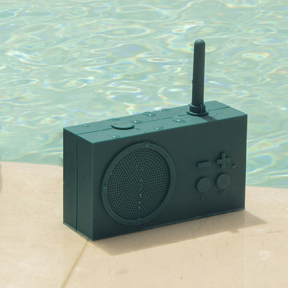 Teal, silicone, rectangular radio with minimal buttons and a short antenna, sitting poolside on a tan stone surface, speckled with water droplets. 