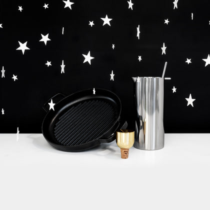 A group of three objects (a cast iron griddle, a stainless steel martini mixer, a gold-colored bottle stopper) on a white surface against a black background with white stars hovering around. 