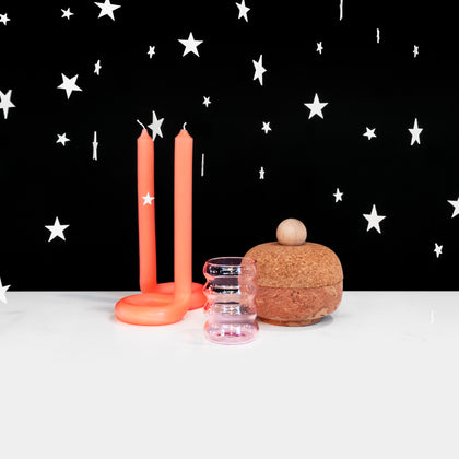 A group of three objects (a neon orange candle with a twisted base, a pink ribbed glass, a round cork container) on a white surface against a black background with white stars hovering around. 
