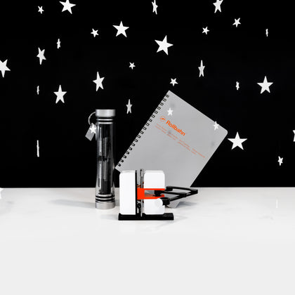 A group of three objects (a glass and metal canister with a digital clock inside, a gray notebook, a square white robotic-looking object with an articulated arm) on a white surface against a black background with white stars hovering around. 