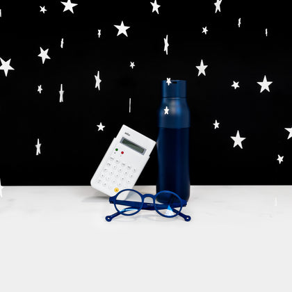 A group of three objects (a navy water bottle, a white calculator, a pair of blue glasses) on a white surface against a black background with white stars hovering around. 