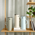 Three cylindrical vacuum jugs in Soft Moss, Sand, and Sky Blue, perch on a wooden bookshelf. Textured off-white curtain in the background.