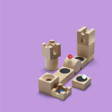 On a purple background, a marble set-up comprised of carved wooden cubes, cylinders, and mini trampolines, with three marbles in red, white, and blue.