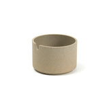 A cylindrical sugar vessel, the color and texture of pressed sand, on a white background. The vessel has straight sides with a slight indentation near the bottom, and has a u shaped cut out near the top for use as a spoon rest.