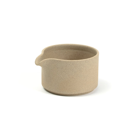 3/4 view of a creamer vessel on a white background. The color and texture are like pressed sand. Creamer has straight sides almost as tall as it is wide. There is a small spout that curves down from the top edge.