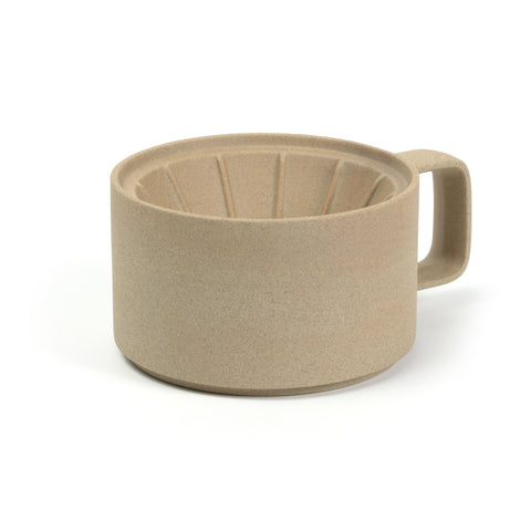 3/4 view of a coffee dripper on a white background. The color and texture is like pressed sand. Dripper has straight sides and a rectangular handle that has rounded corners. The inside of the dripper is a funnel with ridges.