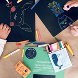 A cropped image of children's hands drawing colorful pictures on sheets of black reusable paper. Wishy Washy colored markers and Butterstix colored chalk are strewn about.