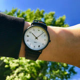 Lifestyle photo of a wrist wearing the Onomatopoeia Watch held up against a vibrant blue sky and green tree in the background.