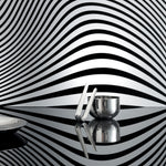 Side view of the sugar bowl with tongs leaning against it, on a flat, reflective surface, the wall behind them is black and white striped with a funhouse curviness. The edge of a dish cuts off on the left side. The dishes reflect each other and the striped background.