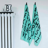 Two light turquoise alphabet tea towels hanging next to each other on a white wall next to a white radiator.