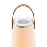 Top portion of a lit Uma Sound Lantern showing the  warm glow of the conical lamp, round flat top with inset circular speaker and volume control rim, outer silver rim dimmer  control and U-shaped wooden carrying handle.