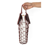 An outstretched hand holds the handles of a small laser cut wine-colored leather net bag, with a white wine bottle inside, its weight stretching the shape of the bag 