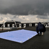 Figure in a rain coat kneeling on a roof to tether down a large blue piece of fabric, with a stormy sky above.