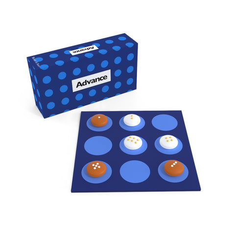 A game in play on a navy square board with nine blue circles arranged in rows of three printed on it. Six disk-shaped games pieces, three white, three brown, each with a different number of raised dots on the top, sit on their own circle. The game box with the same pattern as the board and text “Advance” sits in back.