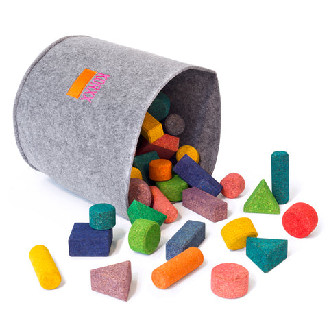 A large grey felt bucket is tipped on its side with cork building blocks tumbling out.  Blocks are multi-colored and multi-shaped. The pink and orange KORXX logo is sewn on the bucket.