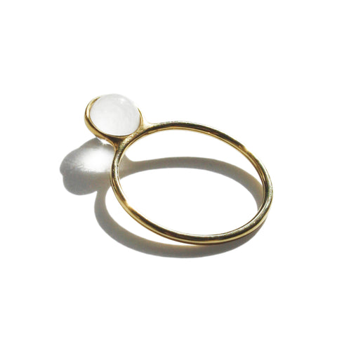 A ring with a gold band lays flat on a white surface.  Extending up from the top of the band is a disc shaped clear smooth stone with a thin gold bezel.  The stone reflects light on the surface below.  