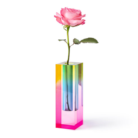 A translucent square block style bud vase emitting a prism of iridescent colors including shades of pink, blue, yellow and green. A single pink rose sits inside.
