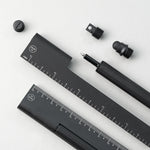 Two views of the Rule One in black and set atop a light gray surface. The bottom view is of the Rule One in its complete set with pen included. The View above this shows the Rule One pen in a dismantled state. Interchangeable tops for the pen are placed around the Rule One. The tops include a stylus, lanyard, and eraser all in black.