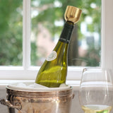 In front of a window, a wine bottle sits on an angle inside a silver ice bucket. It is capped with a Mass Wine Stopper. A glass of wine stands next to the bucket.