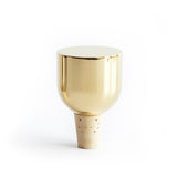 A close up photograph of a Mass Wine Stopper standing cork side down on a white surface.  Sitting on top of the tapered cork, is an inverted shiny brass dome.