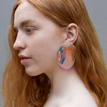 Lifestyle photo of the face and neck of person in profile against a gray background, with long auburn hair, wearing a single Prism Reverberation Earring, an iridescent, slightly irregularly shaped transparent oval shape.