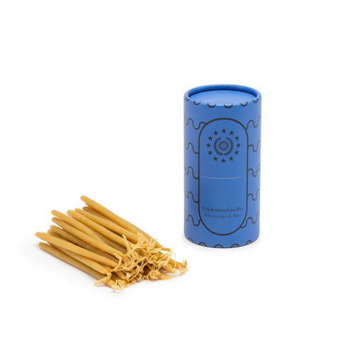 Image featuring a standing blue colored cylinder with black linear wave details along its sides along with the company name and logo, as the container for a set of small mustard yellow candle sticks laying down flat.