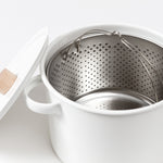 Side view of an open white enamel Kaico Pasta Pan and partial view of round white lid with natural wood knob, and perforated steel strainer inset with folding wire handles