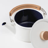 Overhead view of a left facing, open-lid Kaico Kettle on a white background showing the white cylindrical exterior pot and angled pour spout, black interior, folded down wooden topped handle and partially visible white flat-topped lid with wooden knob propped against the kettle’s right side.  