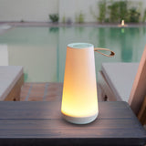 Lit Uma Mini on a wooden table surface in a contemporary, serene setting with a turquoise pool, country tiled patio, cushioned wooden patio furniture and additional Uma Mini located across the pool, each casting soft yellow glow from within the tapered, conical body with a rounded speaker base, flat speaker/dimmer top and handy looped leather side grip.