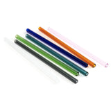 On an angle, six  glass straws in dark grey, green, turquoise, dark blue, orange, and pink lay  flat on a white surface.