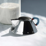 Mirror polished creamer sitting at the base of a glass of milk, against a dappled white background.