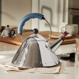 Mirror-polished stainless steel kettle sitting on some papers on a countertop, kitchen accoutrements in the background.
