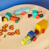 Colorful wooden tractors on a tabletop amongst pretzel pieces, against a blue background.
