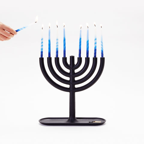 The Menorah is shown with eight burning blue Chanukah candles. A hand reaches out holding the Shammash candle. Used matches are shown in the tray base.