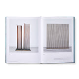 Image of the book opened to reveal inside pages. This spread shows two examples of the artist’s metal sculpture, one on each page. Soldered to metal bases , each piece has multiple thin vertical rods rising upward. 