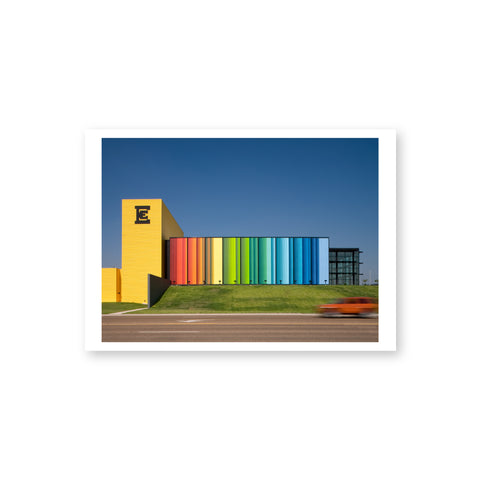 A horizontal postcard with a white border features a daytime exterior view of a contemporary building with a vertical yellow tower, horizontal passageway in rainbow colors red to blue, a square black-framed glass pavilion and a lawn fronted by a street with a moving car.