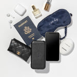 The Carry surrounded by travel accessories: a quilted, black wallet, a US passport, an iPhone, AirPods and Case, a navy, silk eye mask with "i am busy" embroidered on it, and elegantly packaged toiletries.