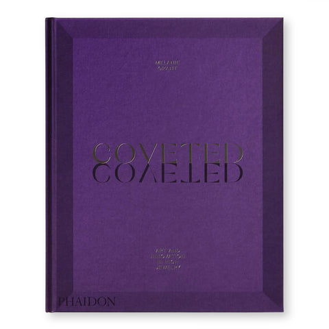 Purple book cover with gold debossed text "COVETED" at center, casting a sharp, mirrored, shadow below.