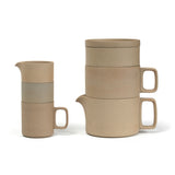 Profile view of two nesting stacks of brown, sandy textured porcelain vessels on a white background. On the left, a cylindrical sugar bowl and creamer fit neatly into a tall mug with a square handle. On the right a minimal tea pot with a small spout and squared handle sits under a cylindrical dripper with a square handle, which is under a soup bowl with a plate on top which can double as a lid. 