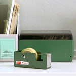 A tabletop deskscape with a dark green, rectangular metal tape dispenser at center, in front of two different file organization boxes.