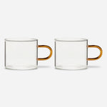 Two identical low glass mugs with straight-sided clear bowls and a delicate, elongated, amber handles.