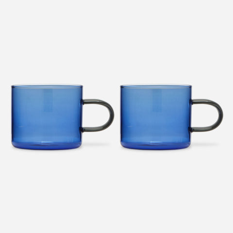 Two identical low glass mugs with straight-sided blue bowls and a delicate, elongated, smoke-gray handles.