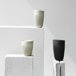 A tiered arrangement of three reusable Huskee Cups on white, plinth-like pedestals, one medium white, one large white and one large black, each featuring flat bottoms, tapering shapes and overall patterns of tight vertical ribs, with a flat, wedge-shaped lids.