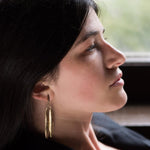 Profile view of a woman wearing the Yew Leaf Earrings. She is leaned back and her chin is jutted up, he earrings hang to her coat collar. The Image is cropped blow her forehead and at her shoulders, there is a window and ledge out of focus in the background.