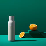 A LARQ Bottle, in granite white, displayed against an emerald backdrop . A halved orange is placed on a tiered stand to the right of the bottle.